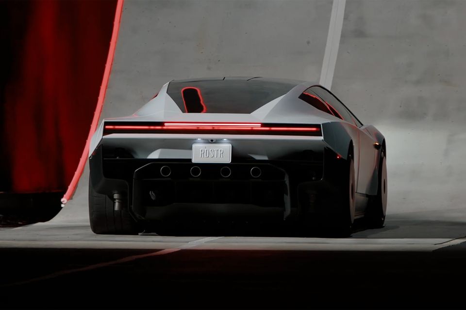 A silver 2026 Tesla Roadster seen from the rear, with a full-width red light bar and quad exhaust-style outlets, parked beside a red curtain