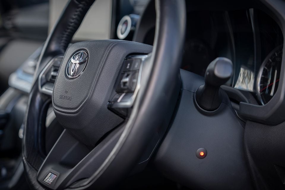 Close-up of a Toyota steering wheel and column stalk inside a vehicle cabin, with steering wheel controls and a small orange indicator light visible on the column