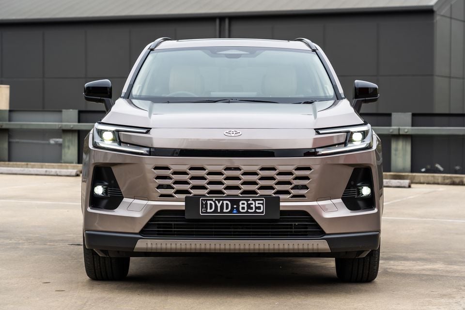Front view of a 2026 Toyota RAV4 Cruiser SUV in a bronze/champagne colour, parked in a car park with headlights on