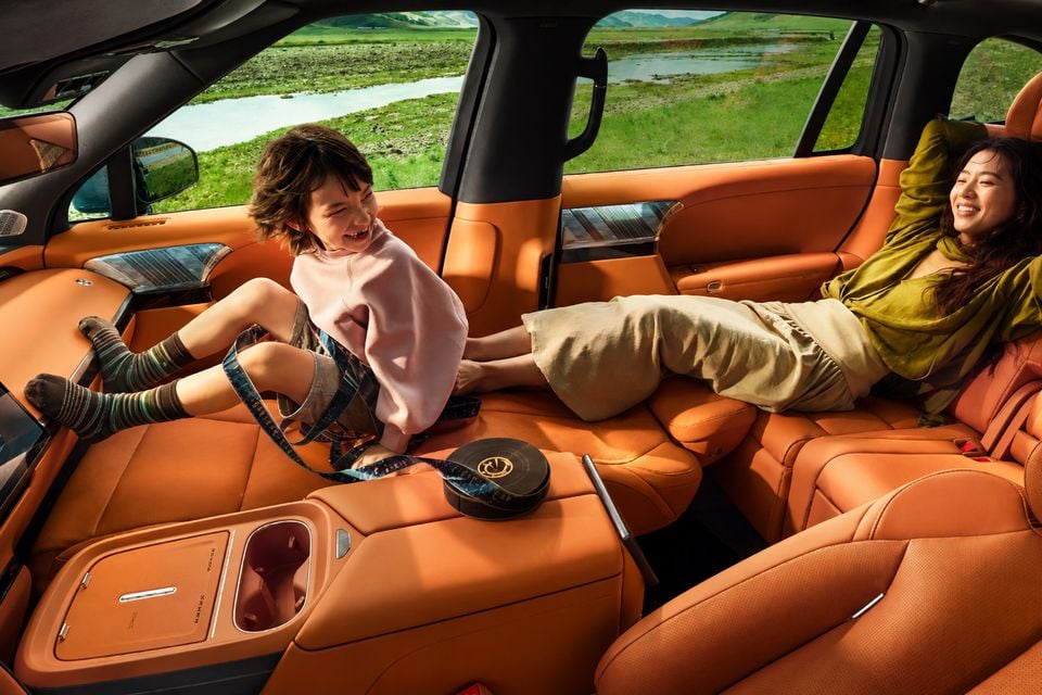 The rear cabin of the 2026 Xpeng G9L SUV showing orange leather seats folded flat, with a child and woman relaxing inside surrounded by a green rural landscape outside.