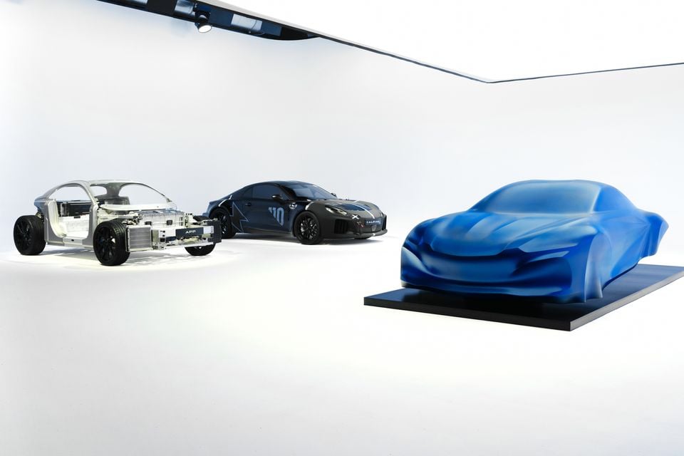 Três estágios do próximo carro esportivo elétrico Alpine A110 são exibidos lado a lado: um chassi vazio, um protótipo camuflado e um modelo coberto por um pano azul revelador.