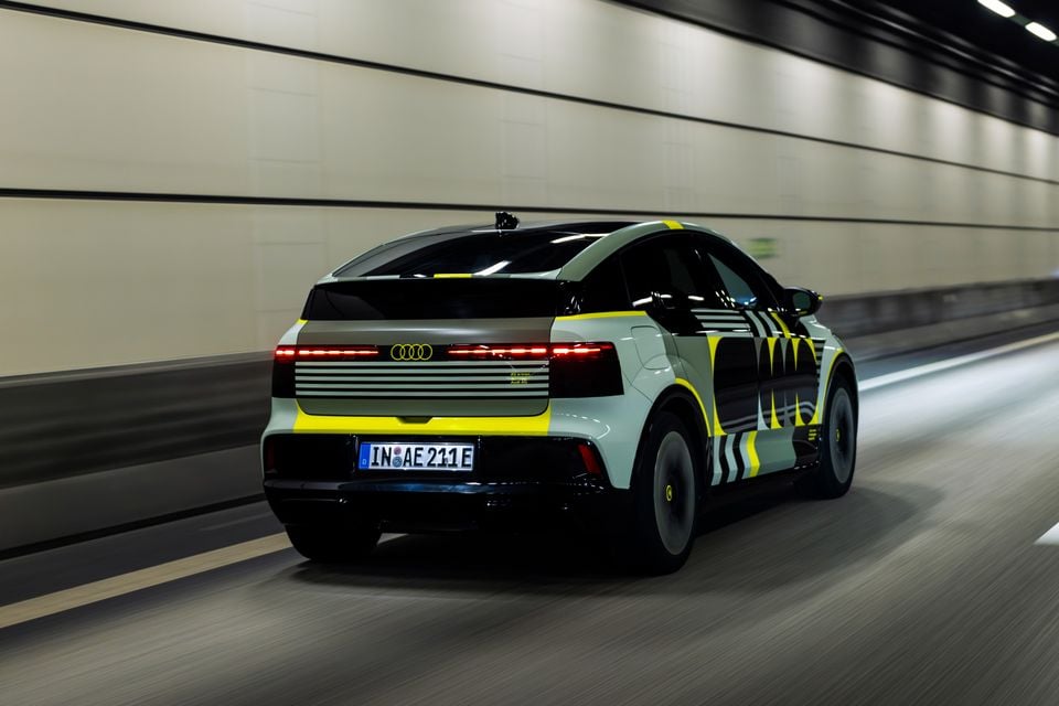 An Audi A2 e-tron prototype with yellow and black camouflage wrap driving through a tunnel, seen from the rear three-quarter angle.