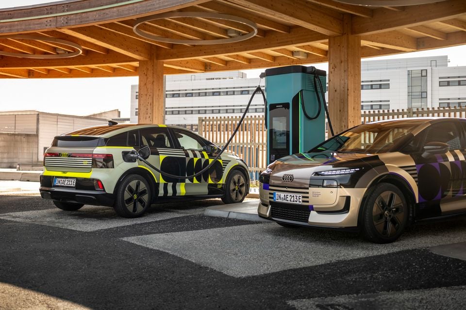 Two Audi A2 e-tron prototypes in camouflage wraps plugged into a Siemens EV charging station under a timber-framed canopy