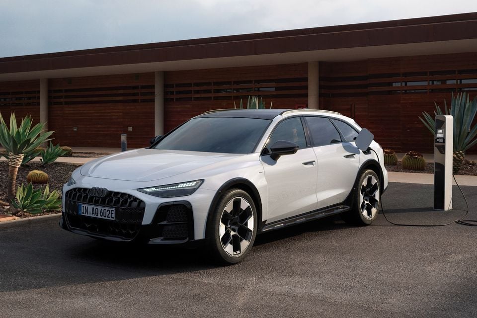 Uma perua Audi A6 Allroad quattro 2027 branca conectada a um carregador de veículo elétrico fora de um prédio revestido de madeira