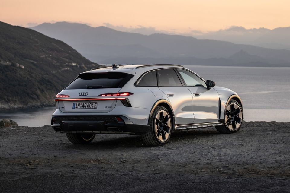 A silver 2027 Audi A6 Allroad quattro wagon seen from the rear, parked on gravel overlooking the sea and mountains at sunset