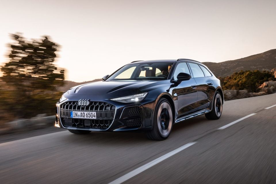 Um Audi A6 Allroad quattro wagon 2027 azul escuro dirigindo ao longo de uma estrada costeira ao pôr do sol, visto de frente