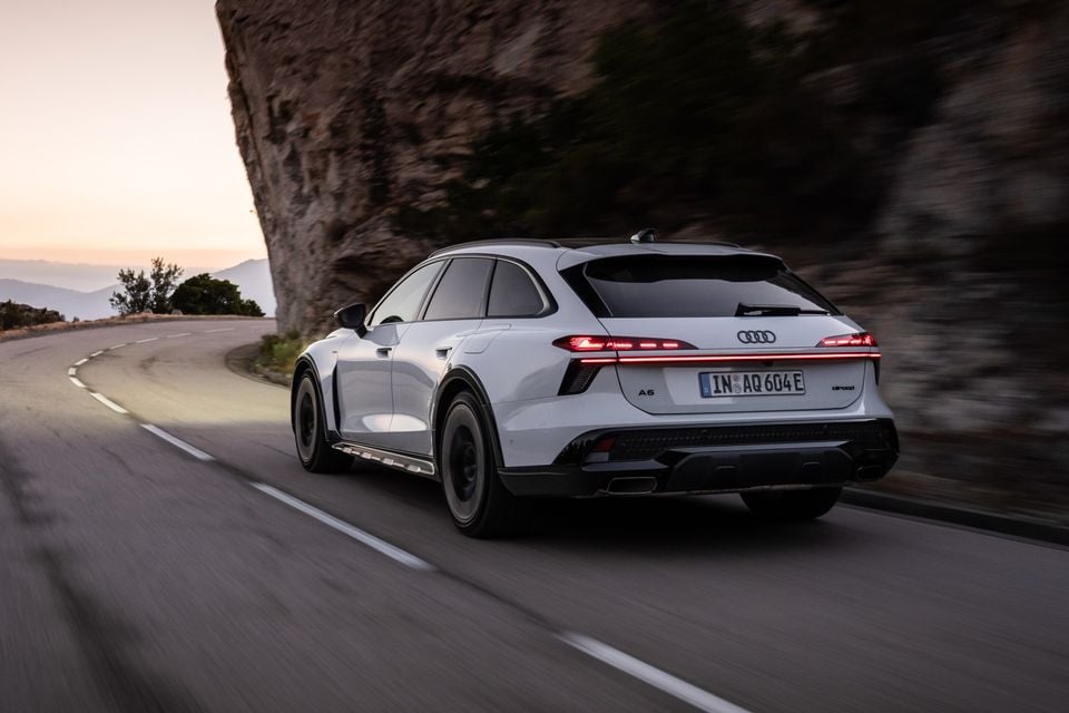 Um Audi A6 allroad quattro wagon branco 2027 dirigindo ao longo de uma estrada sinuosa à beira de um penhasco ao anoitecer, visto de trás