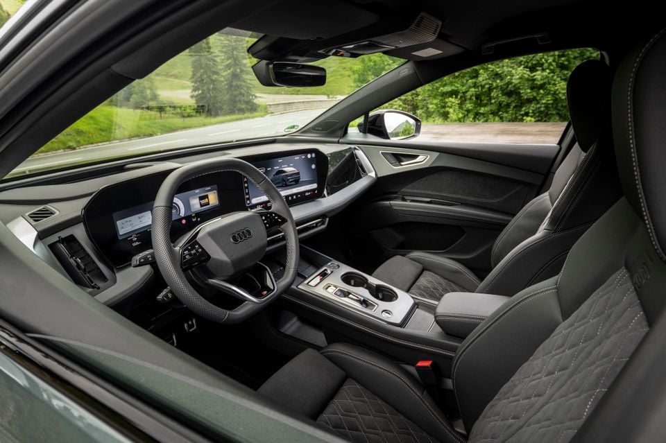Interior of a 2027 Audi Q4 e-tron showing the dashboard, Audi digital display cluster, large infotainment screen, flat-bottomed steering wheel, centre console and quilted dark upholstery seats