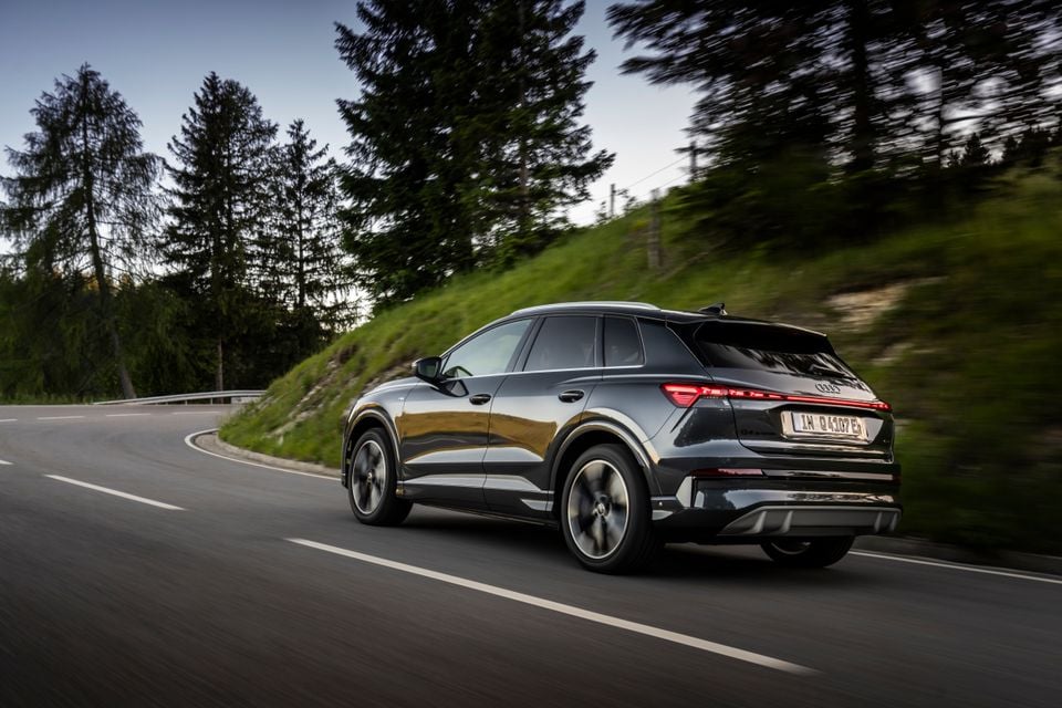 A dark grey Audi Q4 e-tron SUV seen from the rear three-quarter angle, driving along a winding road surrounded by trees
