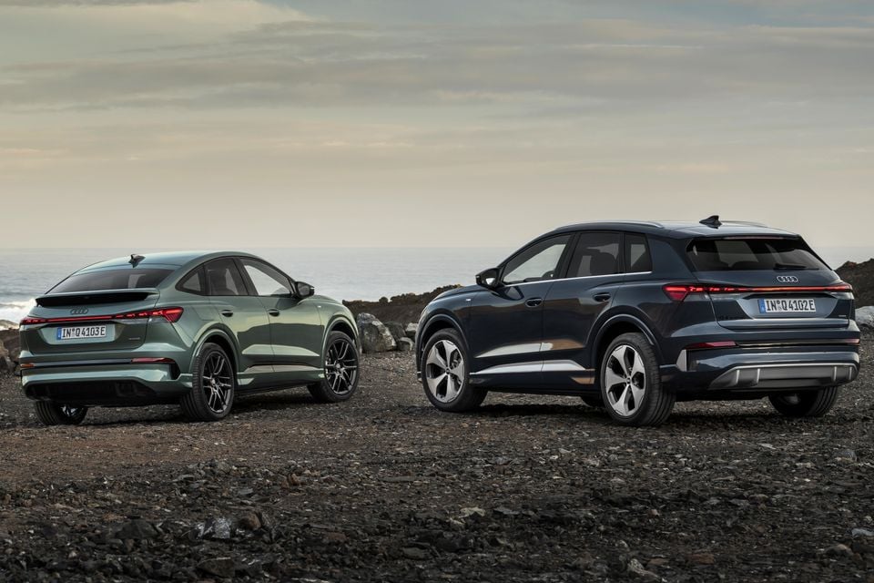 Two Audi Q4 e-tron SUVs seen from the rear in a coastal rocky setting — a green Sportback variant on the left and a dark blue standard SUV on the right
