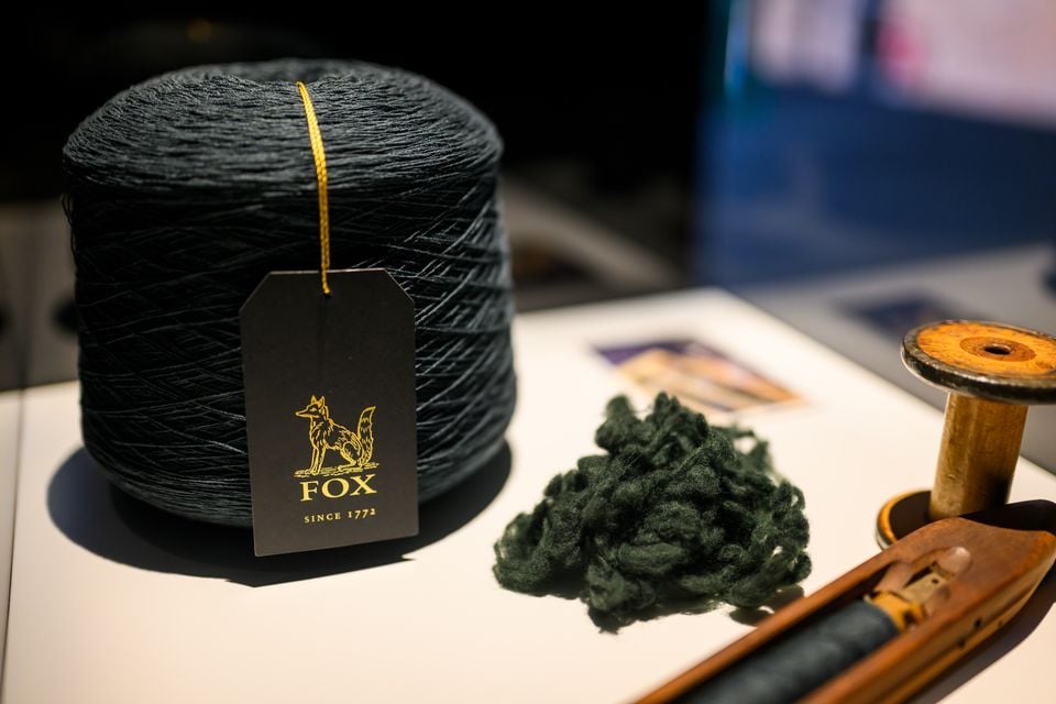 A spool of dark yarn with a Fox Since 1772 branded tag, alongside raw wool fibre and a wooden weaving shuttle, displayed on a white surface