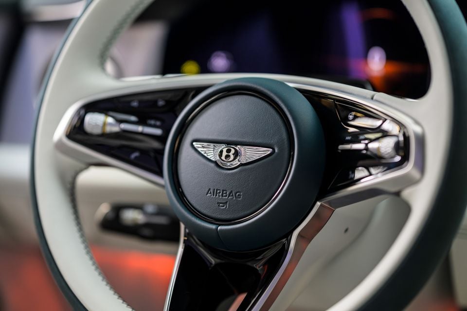Close-up of a Bentley steering wheel with the winged 'B' badge and integrated controls, finished in dark green and cream leather