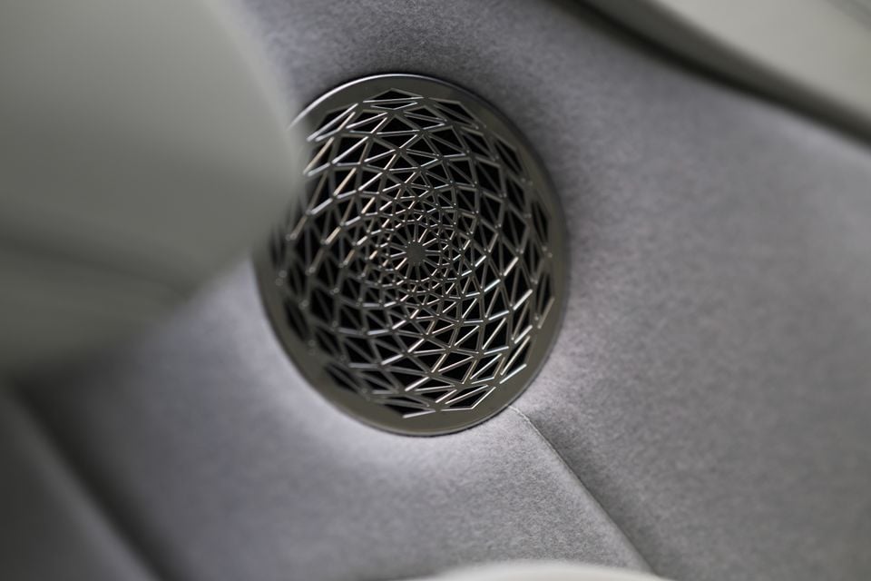 Close-up of a decorative metal speaker grille with an intricate geometric lattice pattern, set into a grey suede-like interior trim panel in a Bentley Torcal