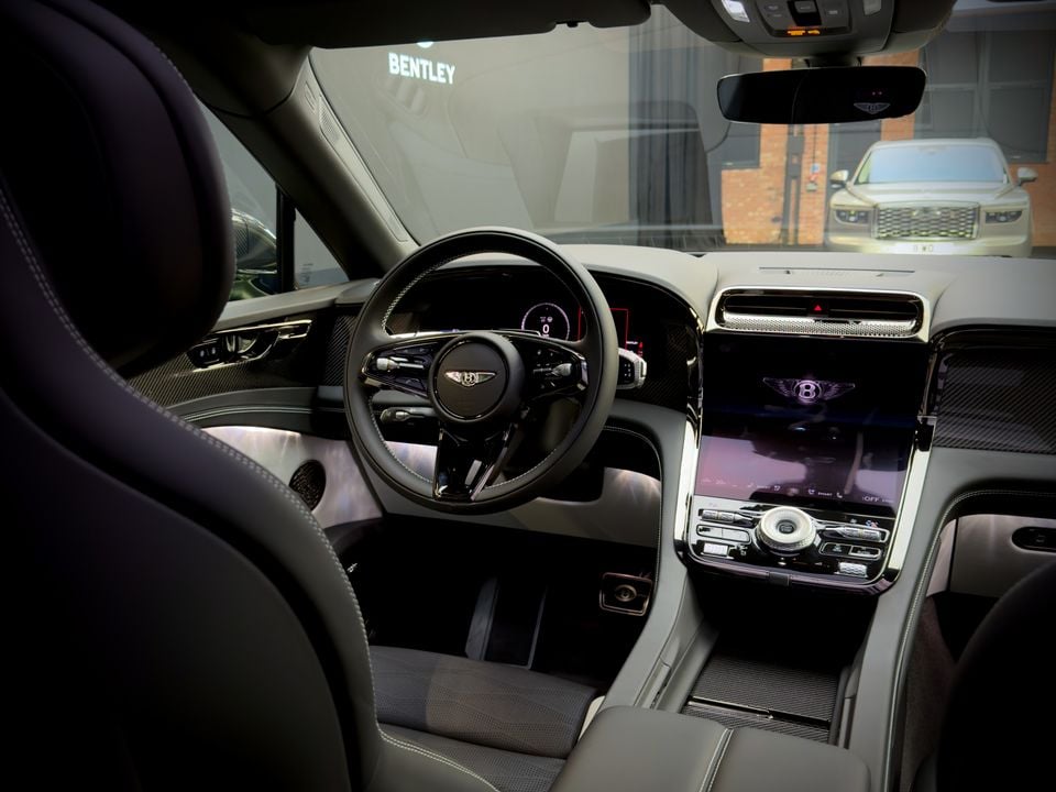 Interior view of a Bentley SUV showing the steering wheel, dashboard and infotainment screen, with another Bentley visible through the windscreen