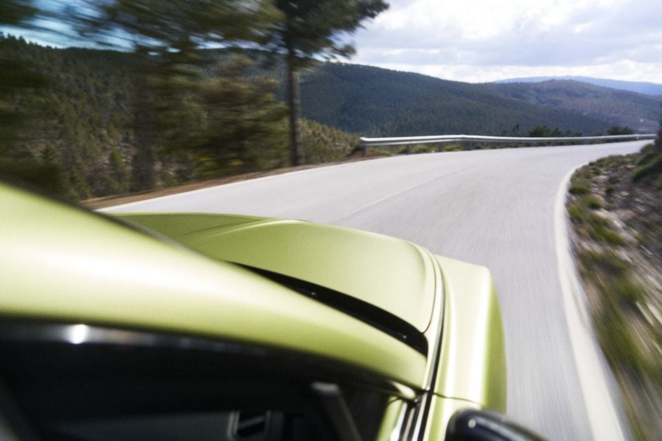 A yellow-green car photographed from the side while driving along a winding mountain road, with forested hills in the background.