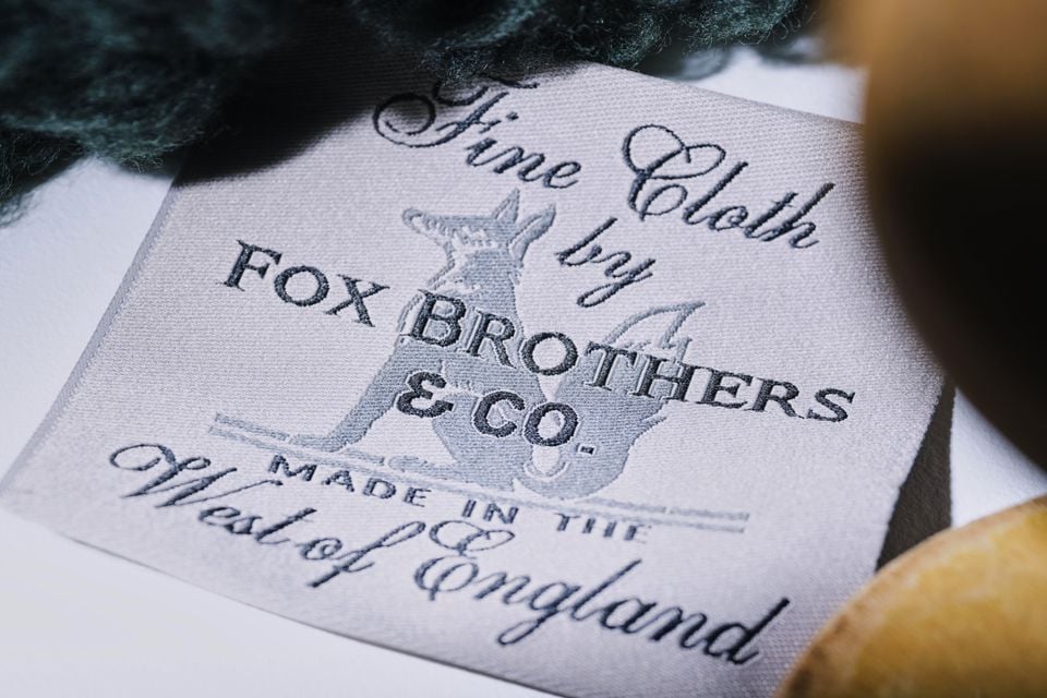 A woven fabric label reading 'Fine Cloth by Fox Brothers & Co., Made in the West of England', with green wool and wooden objects in the background