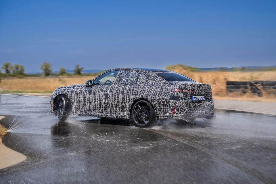 A camouflaged BMW 3 Series prototype sedan seen from the rear three-quarter angle, being driven through a wet handling circuit during testing.