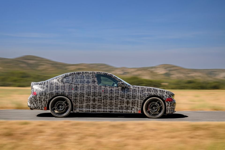 A camouflaged 2027 BMW 3 Series prototype seen from the side while driving on a road through an open landscape
