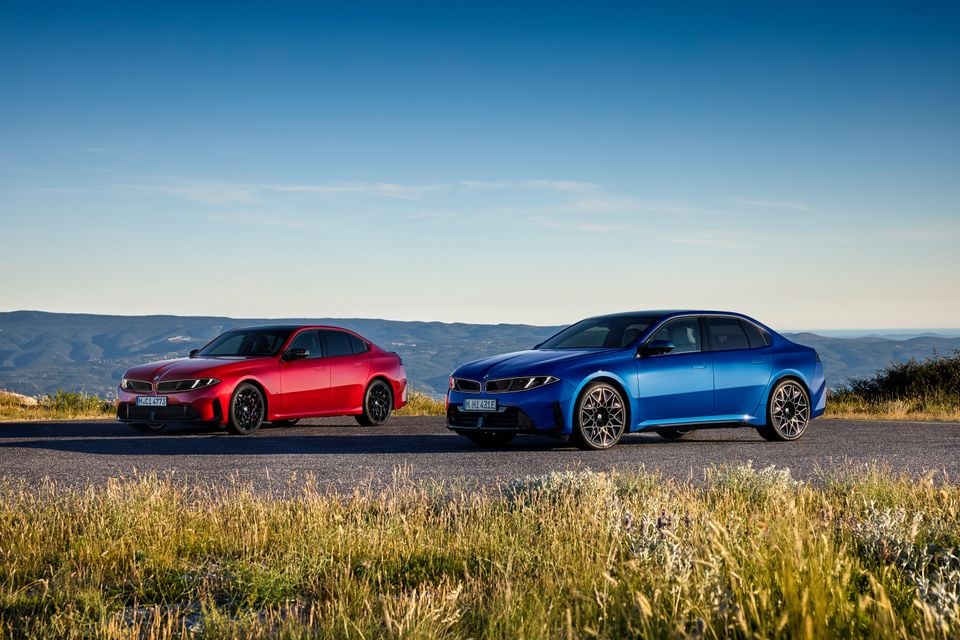 Two 2027 BMW 3 Series sedans, one red and one blue, parked on a mountain road viewed from the front quarter angle.