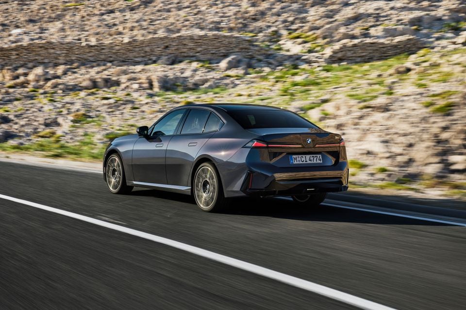A dark grey 2027 BMW 3 Series sedan seen from the rear quarter while driving on a coastal road.