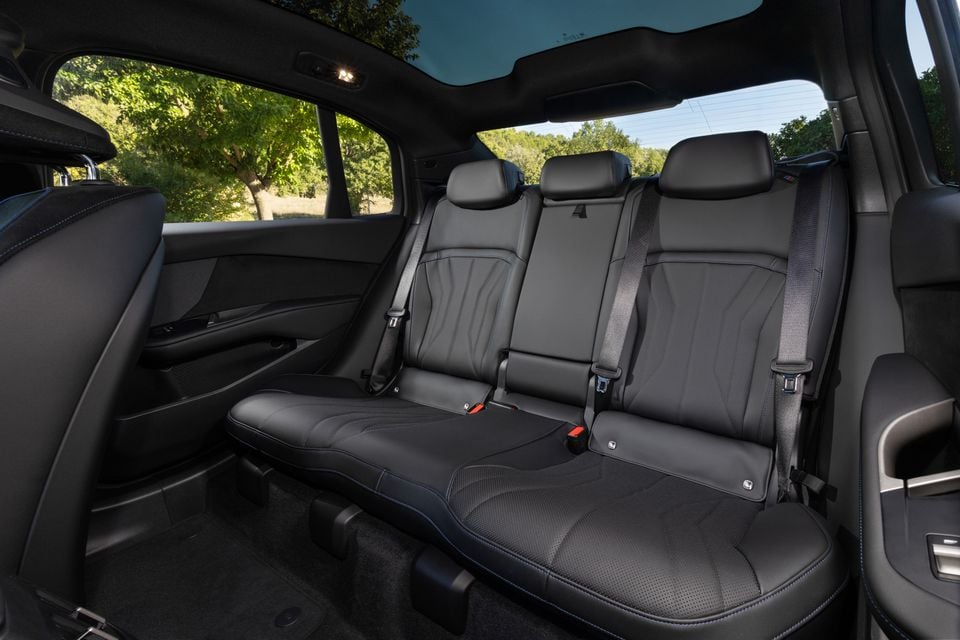 Black leather rear bench seat with blue contrast stitching inside a 2027 BMW i3, seen through the open rear door beneath a panoramic glass roof