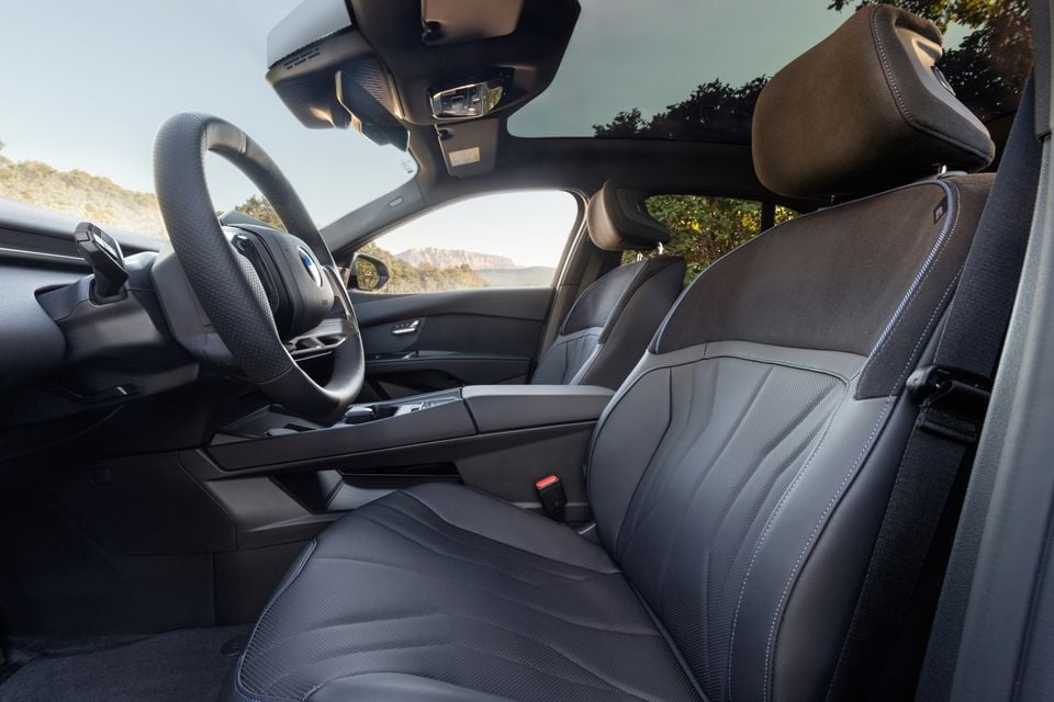 Front seats of the 2027 BMW i3 seen through the driver's door, with grey leather upholstery, steering wheel and panoramic glass roof
