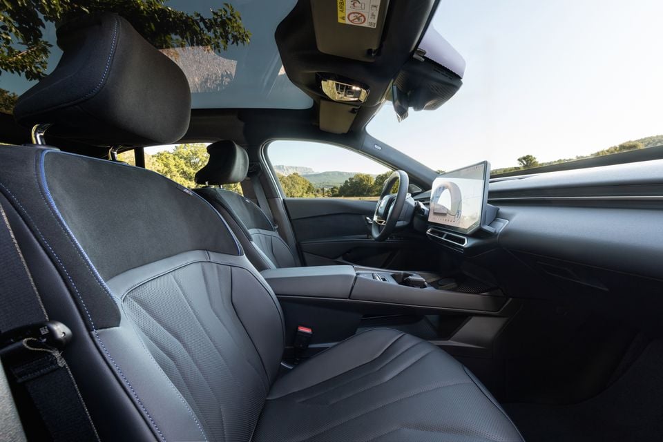 Front seats, steering wheel, centre console and floating touchscreen inside the 2027 BMW i3 50, seen from the passenger side with a panoramic roof