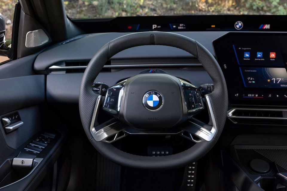 Steering wheel of the 2027 BMW i3 with M badge, panoramic head-up display strip and angled central touchscreen showing climate settings