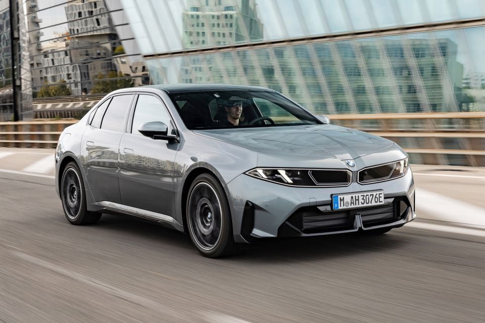 A grey 2027 BMW i3 50 electric sedan driving across a bridge past a glass building, seen from the front.