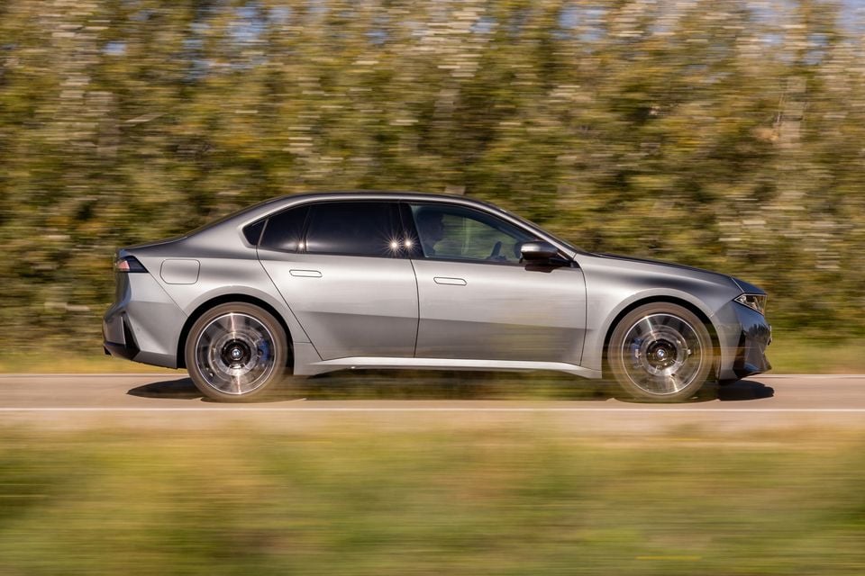 A silver 2027 BMW i3 sedan driving along a country road, seen from the side with motion-blurred trees behind