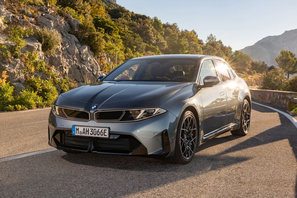 A grey 2027 BMW i3 electric sedan parked on a winding mountain road, seen from the front at an angle