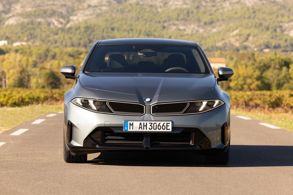 A grey 2027 BMW i3 sedan seen from the front, parked on a country road with vineyards and hills behind