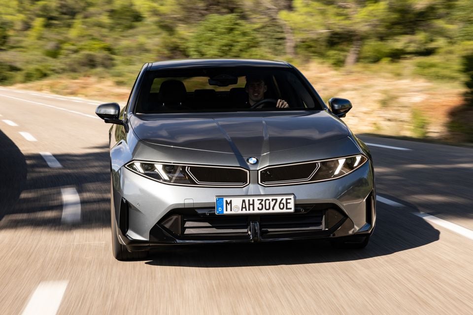 A grey 2027 BMW i3 sedan driving towards the camera on a country road, seen from the front