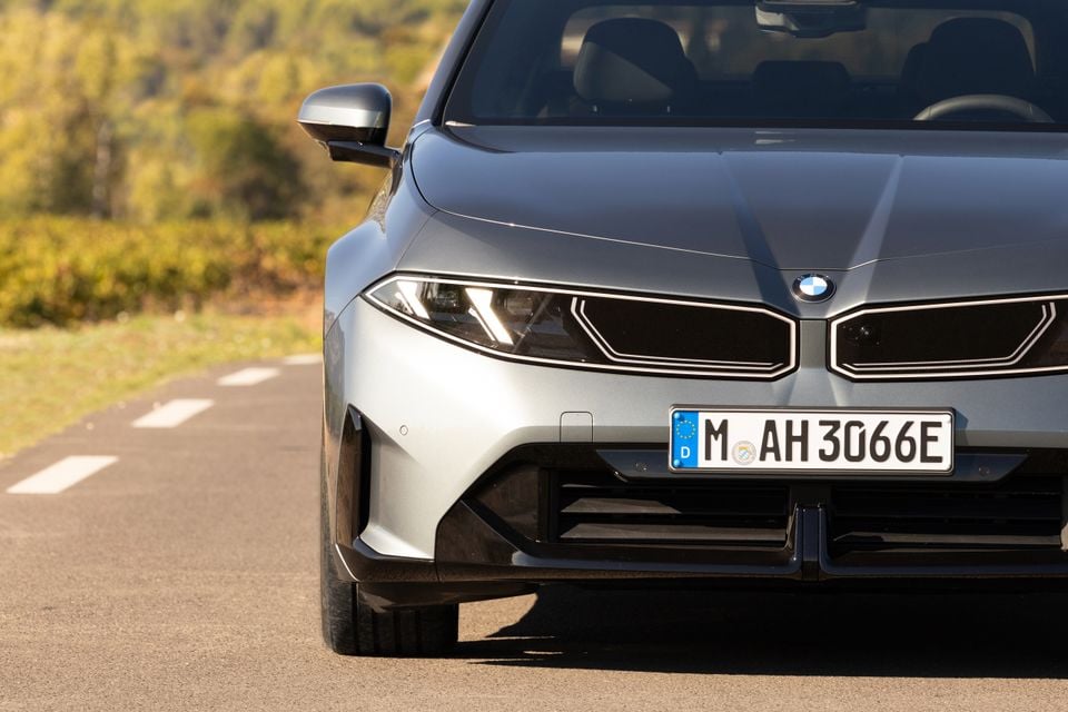 Front of a grey 2027 BMW i3 sedan showing its slim headlights, illuminated kidney grille and badge, parked on a country road