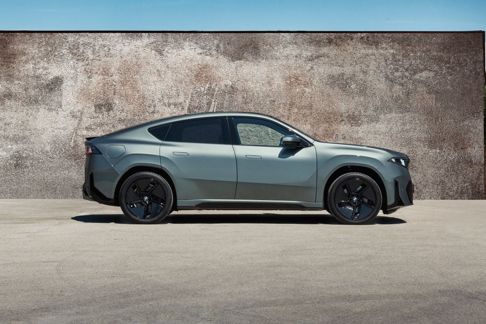 A grey 2027 BMW iX4 50 xDrive SUV coupe photographed side-on against a weathered wall.