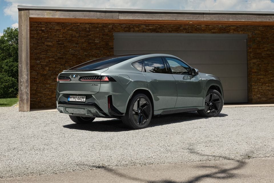A grey 2027 BMW iX4 50 xDrive electric SUV coupe parked on gravel in front of a garage, seen from the rear quarter