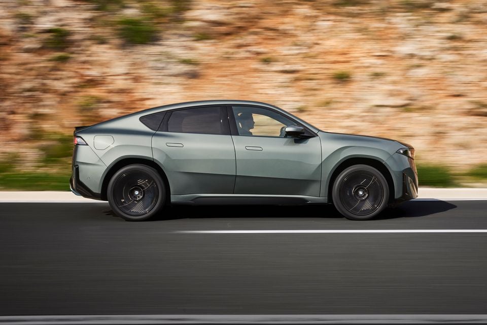 A green 2027 BMW iX4 SUV coupe driving along a road beside a rocky cliff, seen from the side.