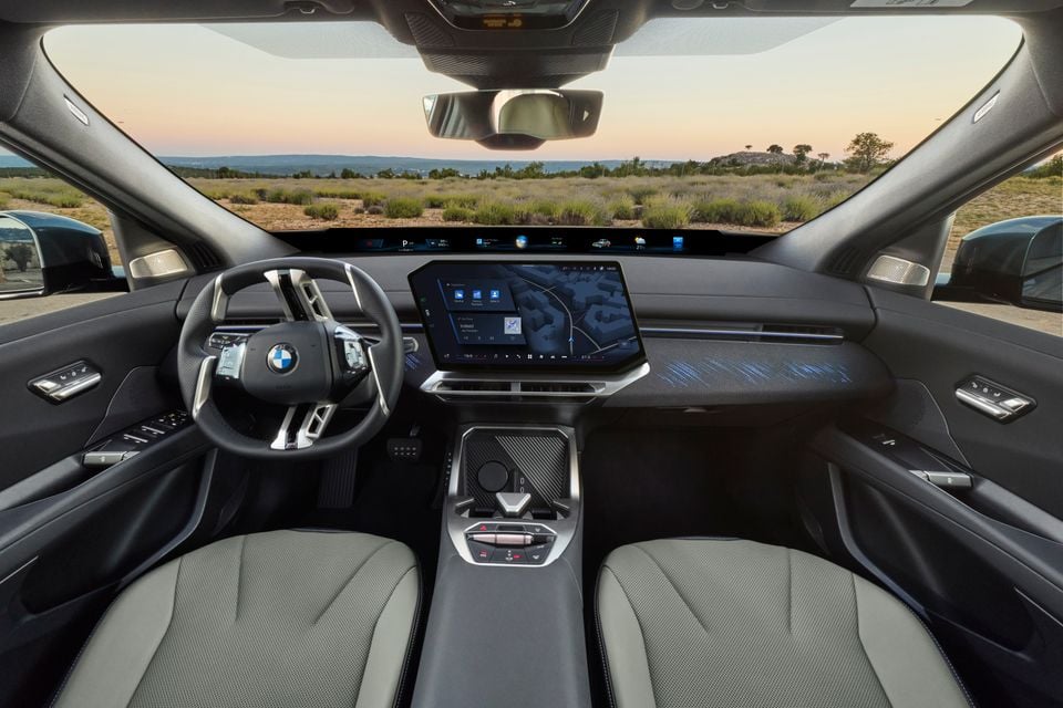 The interior of a 2027 BMW iX4 showing the dashboard, steering wheel, curved touchscreen display and front seats with a desert landscape visible through the windscreen
