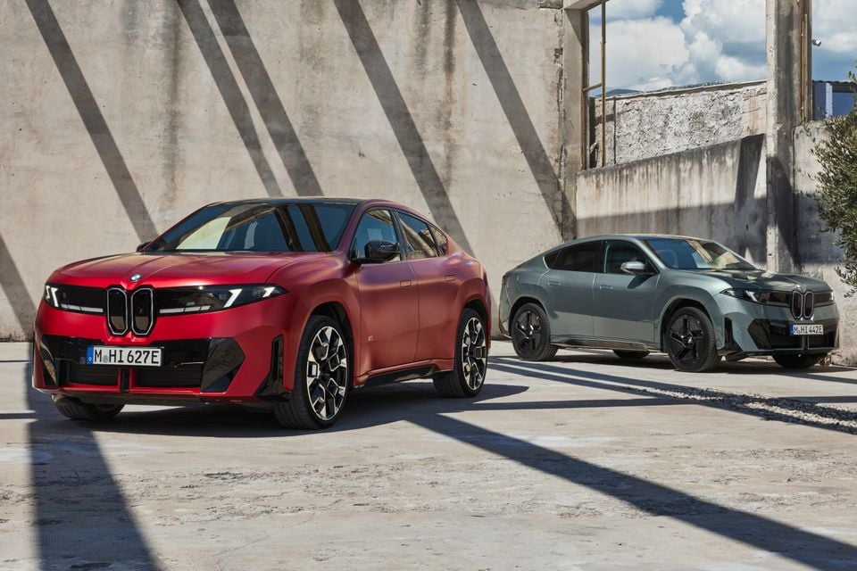 Two 2027 BMW iX4 electric SUVs, one red and one grey, parked facing each other against a concrete wall