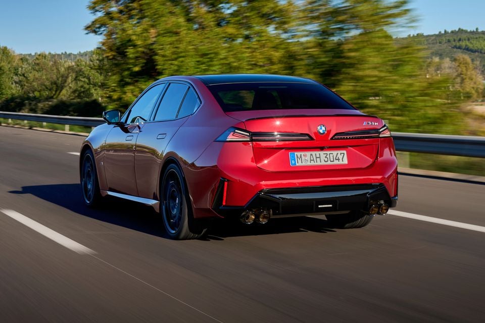 A red 2027 BMW M350 3 Series sedan driving on a highway, seen from the rear, showing its quad exhaust tips