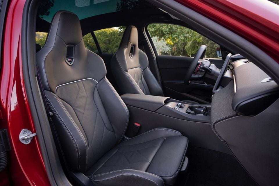 Black leather M sport front seats inside a red 2027 BMW M350 3 Series, seen through the open passenger door with the steering wheel and centre console beyond