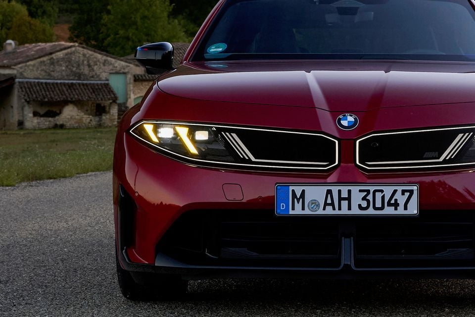 Front end of a red 2027 BMW M350 3 Series with its headlights and illuminated grille glowing, parked on a gravel road at dusk