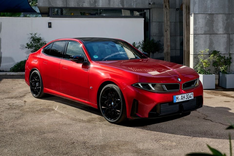 A red 2027 BMW M350 sedan with black wheels parked outside a modern concrete building, seen from the front three-quarter angle
