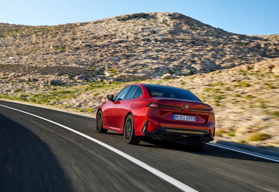 A red 2027 BMW M3 sedan driving on a mountain road, seen from the rear quarter