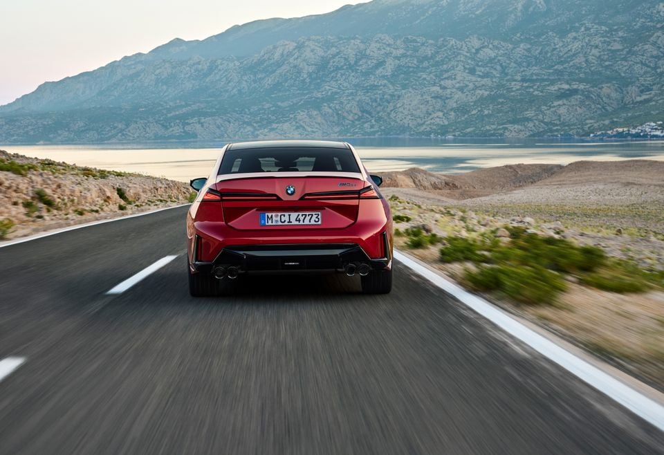 A red BMW M3 sedan drives away from the camera on a coastal road with mountains in the background.
