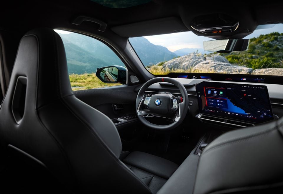 The interior of a 2027 BMW M350 xDrive showing the steering wheel, curved digital display and front sport seats, with mountain scenery visible through the windows.