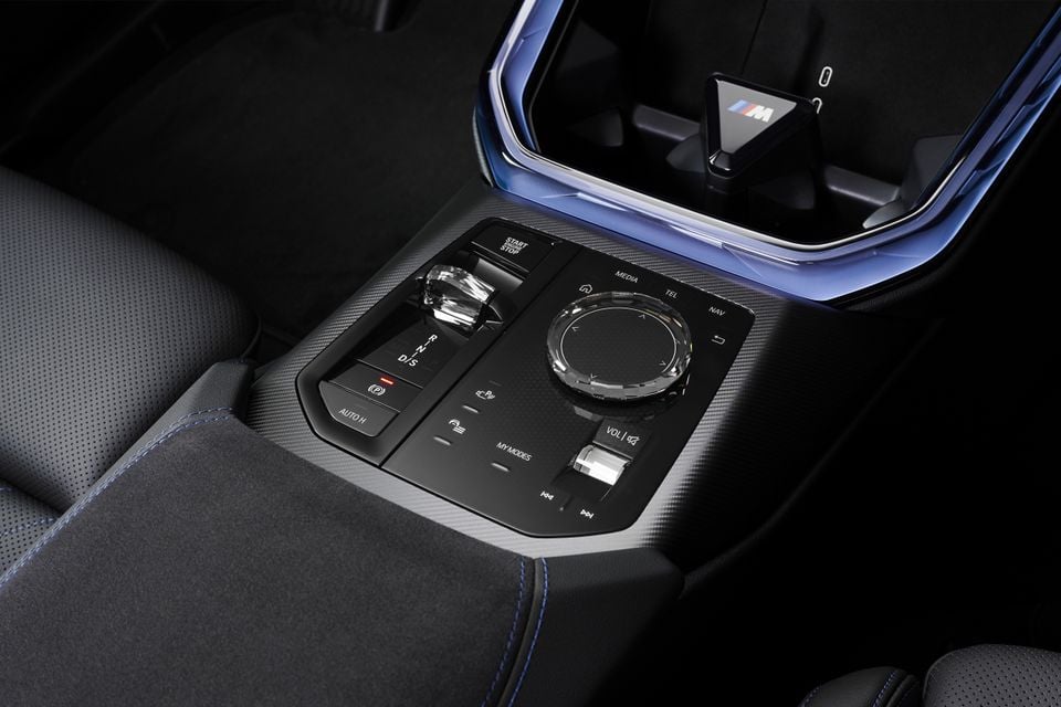 Close-up of the centre console in a 2027 BMW X3 M50, showing the crystal gear selector, iDrive controller, and M badge on the storage tray.