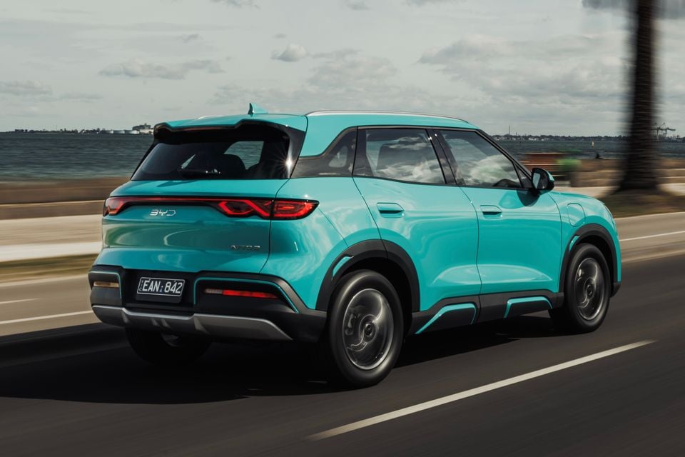 A teal BYD Atto 2 SUV driving along a coastal road, seen from the rear quarter