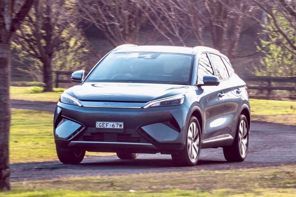 A blue-grey BYD Atto 3 SUV driving along a tree-lined road, seen from the front quarter angle
