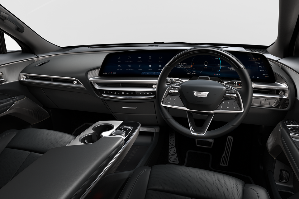 Interior of a Cadillac Lyriq showing the dashboard, curved widescreen display, Cadillac-branded steering wheel, and centre console with cup holders, shot from the passenger side in right-hand drive configuration.
